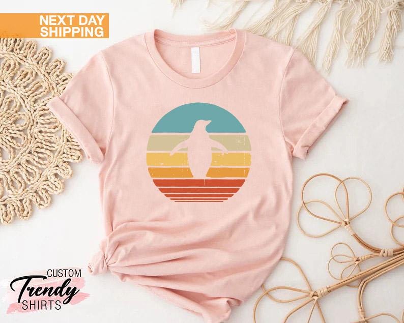 May include: A pink t-shirt with a retro-style graphic of a penguin standing in front of a sunset. The graphic is in shades of blue, yellow, orange, and red.