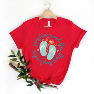 K&ouml;nnte beinhalten: Rotes T-Shirt mit einem Grafikdesign von Flip-Flops, einer Sonne und dem Text "Sunshine, Sand, Flip Flops, Beach Vibes".