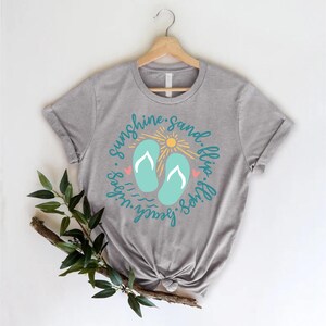 K&ouml;nnte beinhalten: Graues T-Shirt mit einem Grafikdesign von Flip-Flops, einer Sonne und dem Text "Sunshine. Sand. Flip-Flops. Beach. Vibes."