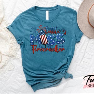 May include: A teal blue t-shirt with a red, white, and blue graphic that says "Little Miss Firecracker". The graphic features a firework with an American flag design.