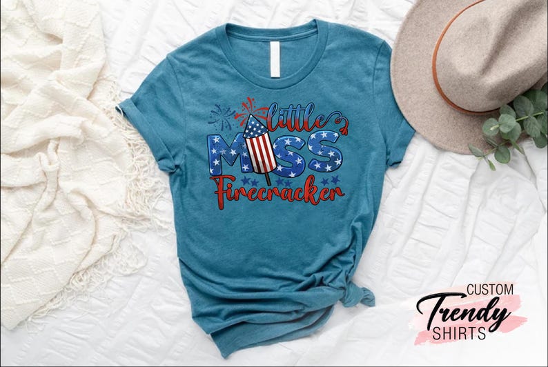 May include: A teal blue t-shirt with a red, white, and blue graphic that says "Little Miss Firecracker". The graphic features a firework with an American flag design.