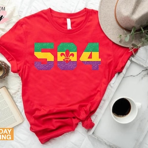 May include: Red t-shirt with a glittery purple, green, and yellow design that says "504" with a fleur-de-lis in the center.