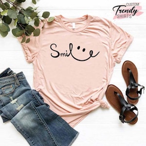 Op de afbeelding: Een lichtroze T-shirt met een zwarte afbeelding waarop "Smile" staat met een smiley.