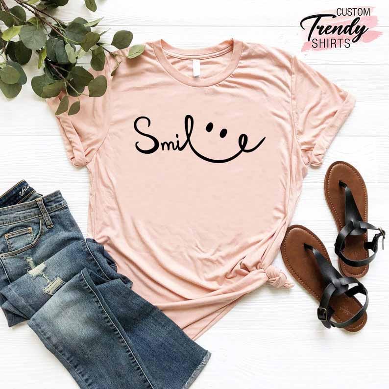 Op de afbeelding: Een lichtroze T-shirt met een zwarte afbeelding waarop "Smile" staat met een smiley.