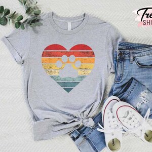 Op de afbeelding: Een grijs T-shirt met een hartvormig ontwerp met een pootafdruk. Het hart heeft een retro kleurenschema van rode, oranje, gele en turquoise strepen. De tekst "CUSTOM Trendy SHIRTS" staat rechtsboven. Het shirt wordt gecombineerd met een blauwe jeans en witte sneakers.