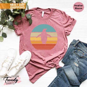 May include: A heather mauve t-shirt with a retro-style graphic of a penguin standing in front of a sunset. The graphic is in shades of blue, yellow, orange, and red.