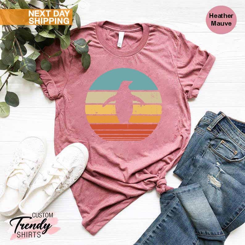 May include: A heather mauve t-shirt with a retro-style graphic of a penguin standing in front of a sunset. The graphic is in shades of blue, yellow, orange, and red.