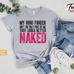 May include: A light gray t-shirt with the text "MY RING FINGER ISN'T THE ONLY PART OF ME THAT LOOKS BETTER NAKED" in black and pink. The shirt is paired with blue jeans and white sneakers. The image also includes a sprig of greenery.
