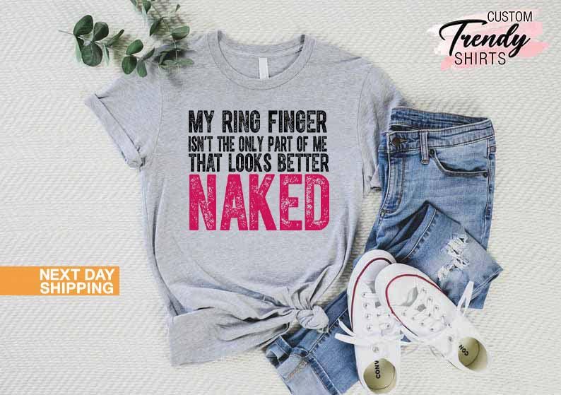 May include: A light gray t-shirt with the text "MY RING FINGER ISN'T THE ONLY PART OF ME THAT LOOKS BETTER NAKED" in black and pink. The shirt is paired with blue jeans and white sneakers. The image also includes a sprig of greenery.