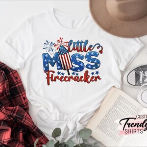 May include: White t-shirt with a red, white, and blue firework graphic and the text "Little Miss Firecracker".