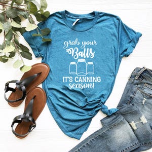 May include: Teal t-shirt with the text "grab your Balls, it's canning season!" in white script. The shirt features a graphic of three mason jars. A pair of black sandals and blue jeans are also in the image.
