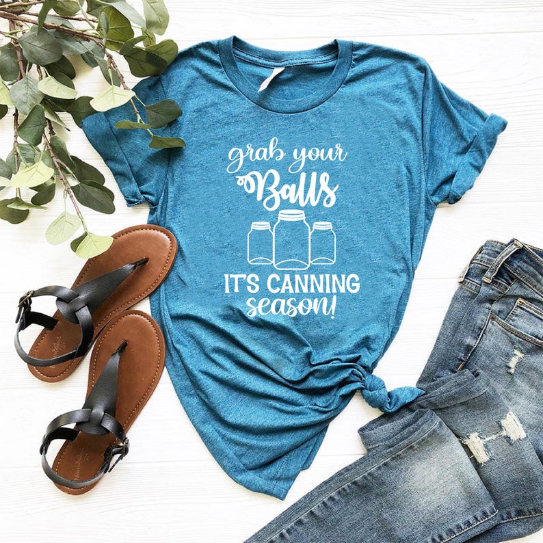 May include: Teal t-shirt with the text "grab your Balls, it's canning season!" in white script. The shirt features a graphic of three mason jars. A pair of black sandals and blue jeans are also in the image.