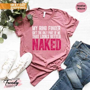 May include: Heather mauve t-shirt with the text "MY RING FINGER ISN'T THE ONLY PART OF ME THAT LOOKS BETTER NAKED" in white and pink. The shirt is paired with blue jeans and white sneakers.