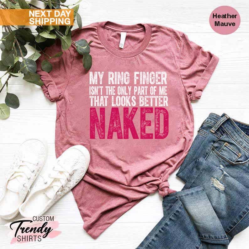May include: Heather mauve t-shirt with the text "MY RING FINGER ISN'T THE ONLY PART OF ME THAT LOOKS BETTER NAKED" in white and pink. The shirt is paired with blue jeans and white sneakers.