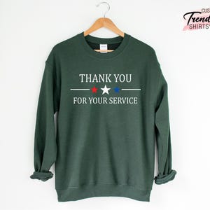 May include: A dark green sweatshirt with the text "THANK YOU FOR YOUR SERVICE" in white, with a red star and a blue star. The sweatshirt is hanging on a wooden hanger.
