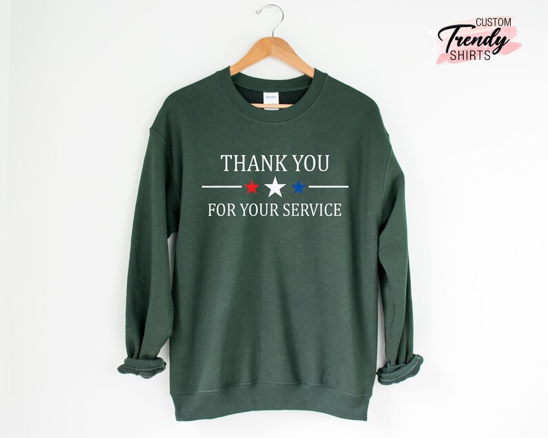 May include: A dark green sweatshirt with the text "THANK YOU FOR YOUR SERVICE" in white, with a red star and a blue star. The sweatshirt is hanging on a wooden hanger.