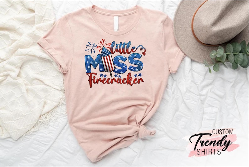 May include: A pink t-shirt with a graphic design featuring a firework with an American flag design and the text "Little Miss Firecracker".