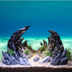 Puede incluir: Decoración de acuario con una disposición naturalista de rocas y madera flotante. La pieza central tiene un aspecto gris oscuro y desgastado con bordes irregulares, complementado con rocas azul-grisáceas más pequeñas y plantas verdes. El fondo sugiere un entorno submarino.