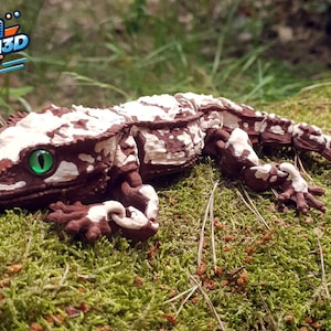 Puede incluir: Una figura de gecko articulada impresa en 3D con un patrón moteado en marrón y blanco por todo el cuerpo. El lagarto tiene una superficie texturizada y un ojo verde brillante con una pupila negra vertical. El logo KOGI3D es visible en la esquina.
