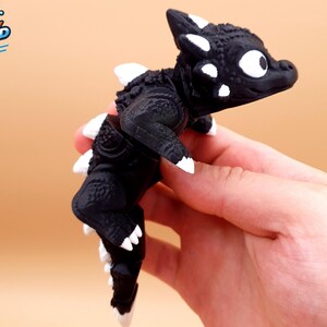 Puede incluir: Una figura de dragón articulada de color negro con detalles blancos en los cuernos, las espinas dorsales, las garras y los ojos. La criatura presenta una superficie texturizada y escamosa con una cabeza ancha y redondeada. El logo KOGI3D es visible.