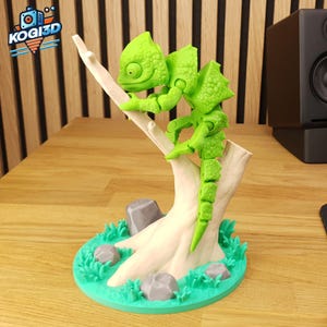Pode incluir: Uma figura impressa em 3D de um camaleão verde articulado em um galho branco. A escultura está em uma base verde com pedras cinzentas e grama. O logotipo "KOGI3D" é visível no canto superior esquerdo.