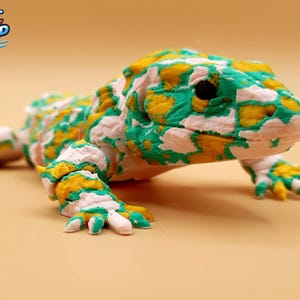 Op de afbeelding: Een 3D-geprinte, beweegbare hagedisfiguur met een gevlekt patroon in wit, heldergroen en goudgeel. De hagedis heeft een textuurrijk oppervlak, een afgeronde kop met zwarte ogen en vier poten met zichtbare gewrichten voor beweging.