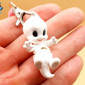 May include: A white ghost keychain featuring a small, articulated figure with black eyes and a smiling mouth. The ghost has raised arms and a pointed tail, attached to a silver-colored metal lobster clasp. The figure has a smooth, matte finish.