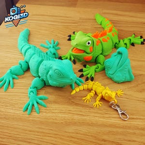 Puede incluir: Una colección de figuras de iguana impresas en 3D en varios tamaños y colores. La más grande es turquesa, la del medio es verde con detalles naranjas y la más pequeña es amarilla con un llavero. El logotipo "KOGI3D" está en la esquina superior izquierda.