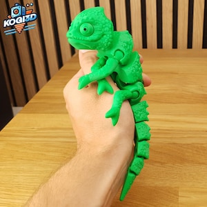 Puede incluir: Un juguete de camaleón verde brillante y articulado. El juguete se sostiene en una mano y presenta un diseño detallado y texturizado. El logotipo "KOGI3D" es visible en la esquina superior izquierda. El cuerpo del camaleón está segmentado.