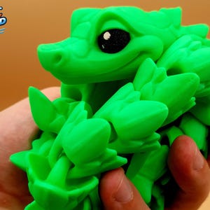 May include: A bright green articulated 3D printed lizard figurine with a textured, leaf-like body and large, dark, glossy eyes. The creature features a segmented design that allows for movement, showcasing a vibrant neon green color throughout its form.