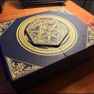 Peut inclure: Une boîte bleu foncé avec des motifs géométriques dorés et des coins décorés. La boîte présente un emblème central doré et un couvercle hexagonal. Le design suggère un thème fantastique ou de jeu.