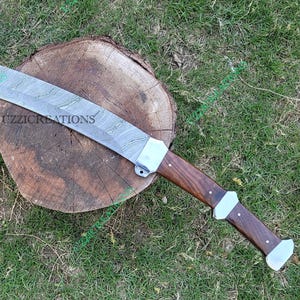 May include: A curved blade sword with a patterned steel blade and a wooden handle. The handle is dark brown with silver-coloured accents. The sword is resting on a wooden tree stump, set against a backdrop of green grass.
