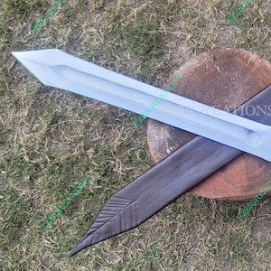 May include: A silver-bladed sword with a wooden handle and a leather sheath. The handle is intricately carved, and the sheath is dark brown with decorative stitching. The sword rests on a wooden surface.