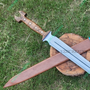 May include: Two crossed swords with sheaths rest on a wooden surface surrounded by grass. One sword has a light blue blade and a brown handle. The other sword has a brown sheath with a blue and red handle. The text "UZZICREATIONS" is visible.