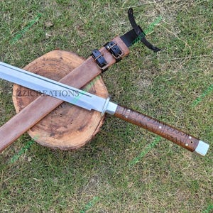 May include: A silver-bladed sword with a brown, textured handle and a matching sheath. The sword rests on a wooden surface, with the sheath and handle detailed with leather and metal accents. The text "22ZICREATIONS" is visible on the blade.