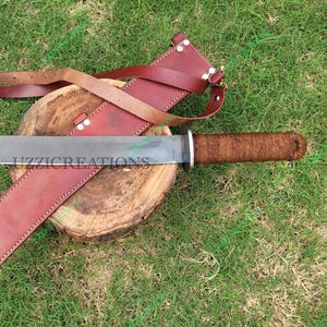 May include: A large knife with a silver blade and a brown, wrapped handle. It is accompanied by a red leather sheath and strap. The knife rests on a wooden surface, with green grass in the background. The text "UZZICREATIONS" is visible on the blade.