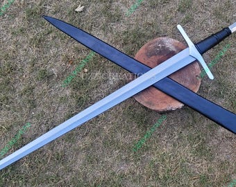 May include: A silver-colored sword with a cross-shaped hilt and a black handle rests atop a black scabbard. The sword and scabbard are positioned on a wooden surface, with a grassy background.