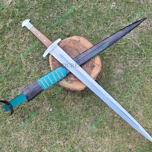 May include: A medieval sword with a silver blade and a wooden handle, lying on a wooden surface. The sword is paired with a black sheath with teal details. The image is taken outdoors on grass.