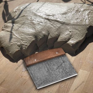 May include: A stainless steel dough scraper with a wooden handle. The scraper has a textured blade and is sitting on a wooden surface.