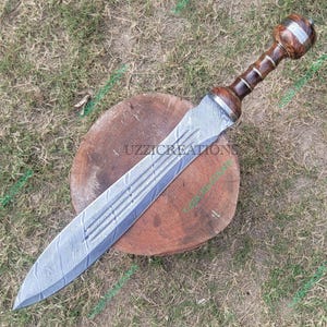 May include: A handcrafted gladiator sword with a wooden handle and a silver blade. The handle is a dark brown colour with silver accents. The blade has a textured pattern. The sword is resting on a wooden surface.
