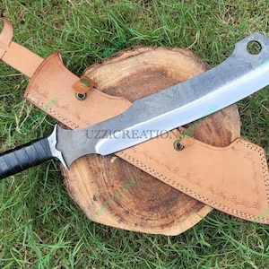 May include: A large, silver-colored cleaver-style knife with a black handle and a leather sheath. The knife rests on a wooden surface, with the sheath partially covering it. The background is green grass. The text "UZZICREATION" is visible.