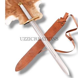 May include: A medieval sword with a silver blade, gold crossguard, and a white and silver wrapped handle. It is displayed with a brown leather sheath and strap. The sword rests on a brown fur-like surface. The text "UZZICREATIONS" is visible.