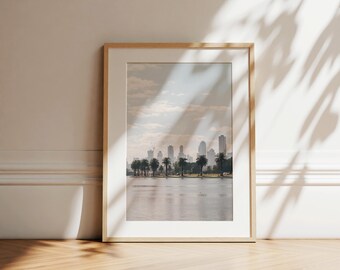 May include: A framed art print featuring a serene cityscape with a body of water in the foreground. The image showcases a skyline of tall buildings, palm trees, and a cloudy sky. The frame is a light wood color, adding a natural touch to the artwork.