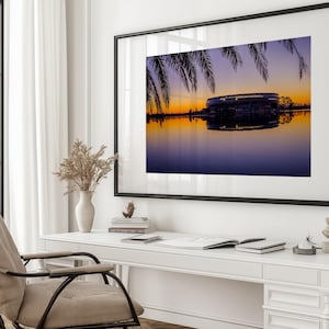 May include: A framed art print depicting a sunset over water, a building silhouette, and palm fronds. The artwork hangs above a white desk with a chair, books, and decorative items, creating a serene office setting.