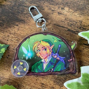Peut inclure: Un porte-clés en acrylique transparent représentant un personnage aux cheveux blonds et aux oreilles pointues portant une tunique verte, accompagné d'une fée bleue lumineuse. Le motif est encadré par une bordure brune en forme d'arbre.