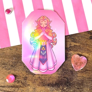 Peut inclure: Un autocollant rectangulaire aux coins coupés présentant une illustration stylisée d'un personnage elfe blond aux oreilles pointues, portant une robe rose avec un corsage doré et un emblème Triforce, sur un fond rose doux et holographique.