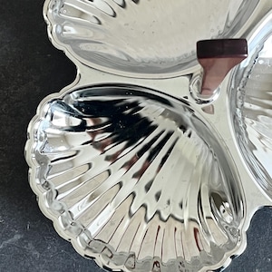 May include: A silver-colored, three-section serving dish with a shell-like design. The dish has a dark brown handle and a scalloped edge. The dish is made of a shiny, reflective material.