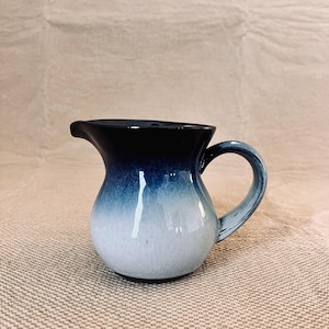 Op de afbeelding: Een keramische kan met een afgeronde basis en een handvat. De kan heeft een kleurovergang, die overgaat van een donker marineblauw aan de bovenkant naar een lichtblauw en wit aan de onderkant. De kan heeft een tuit om uit te schenken.