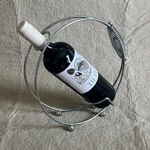 May include: A silver metal wine bottle holder with a circular design. It holds a dark wine bottle with a white label. The holder has a spring-like handle and rests on three small silver spheres.