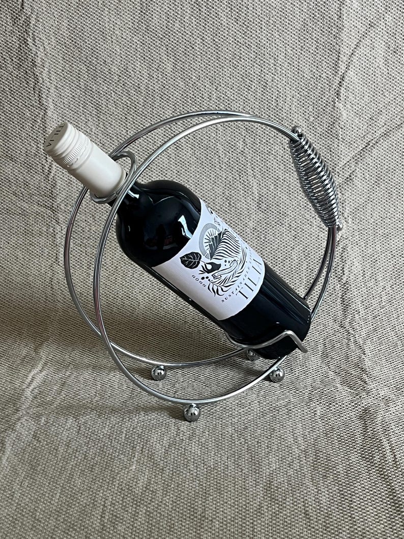 May include: A silver metal wine bottle holder with a circular design. It holds a dark wine bottle with a white label. The holder has a spring-like handle and rests on three small silver spheres.